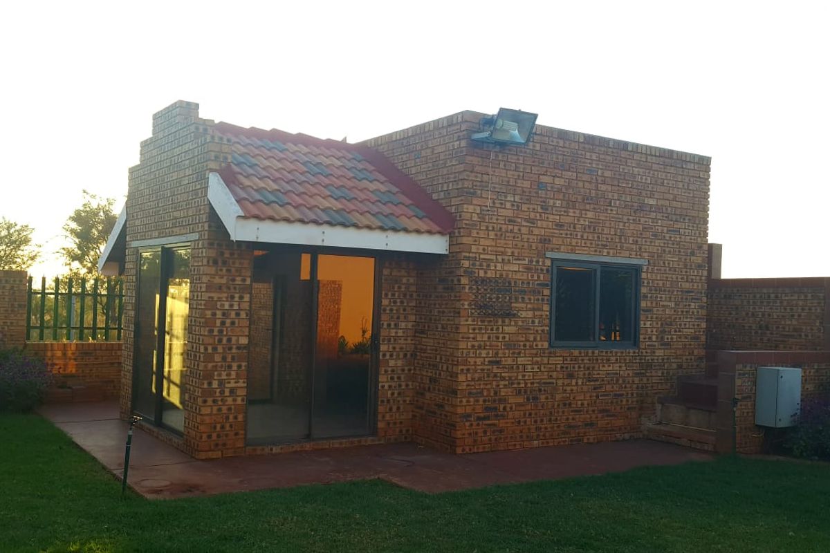 A rear view of a face brick house featuring a tiled roof section, sliding glass doors, and a paved patio area set within a garden.