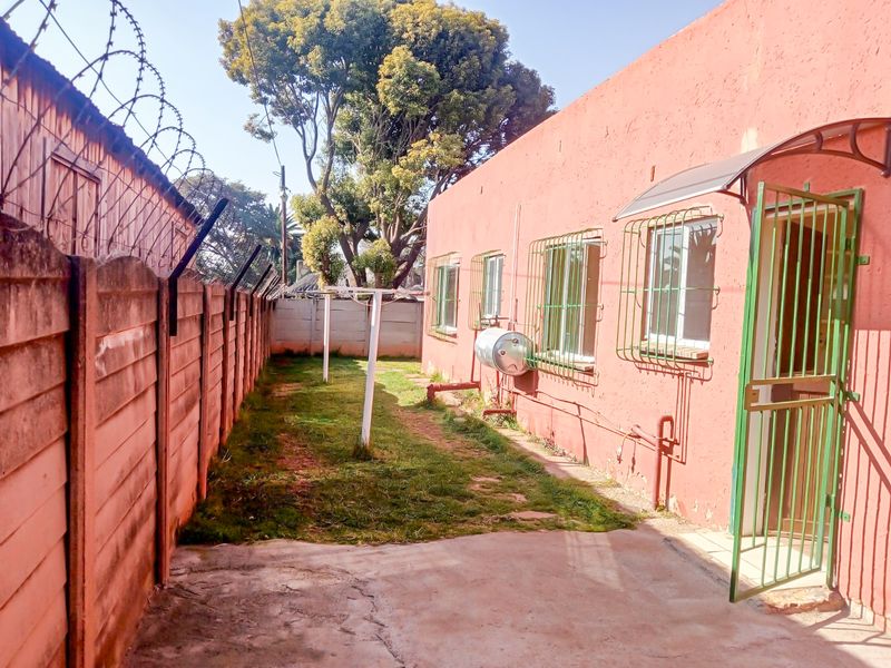 Rear exterior view of the cottage featuring a concrete boundary wall with security fencing, paved area, and external geyser unit.
