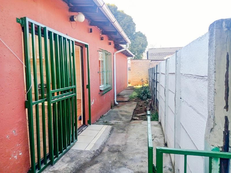 A side exterior view of the cottage featuring a concrete pathway, security gates, and a precast concrete boundary wall.