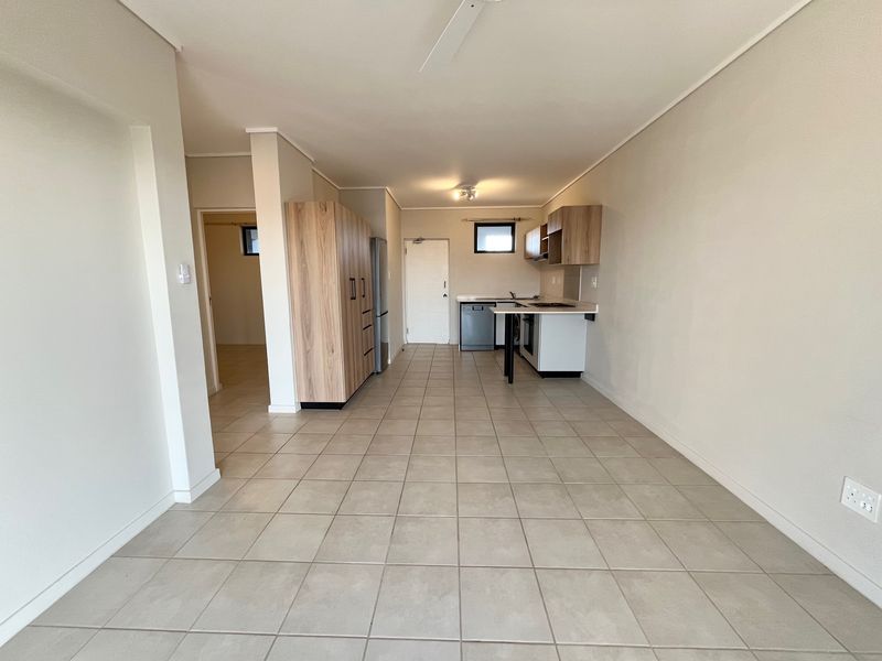 An open-plan living and kitchen area featuring neutral tiled flooring, built-in cabinetry, and integrated kitchen appliances in a clean, vacant space.