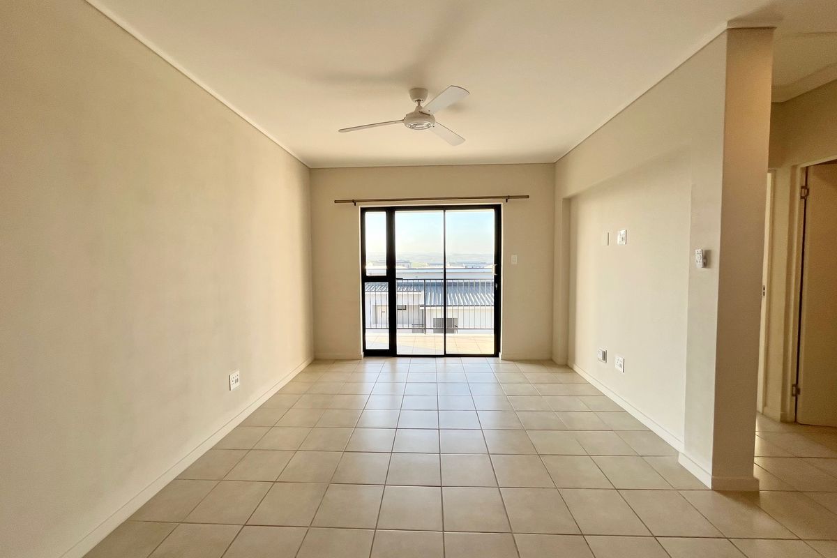 An empty, well-lit living area featuring neutral ceramic floor tiles, a ceiling fan, and a sliding glass door leading to an outdoor balcony.