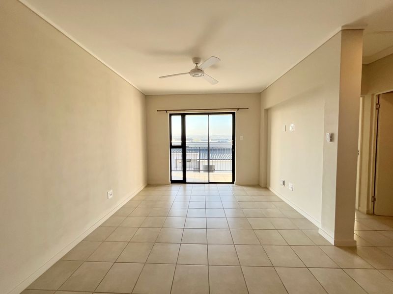 An empty, well-lit living area featuring neutral ceramic floor tiles, a ceiling fan, and a sliding glass door leading to an outdoor balcony.