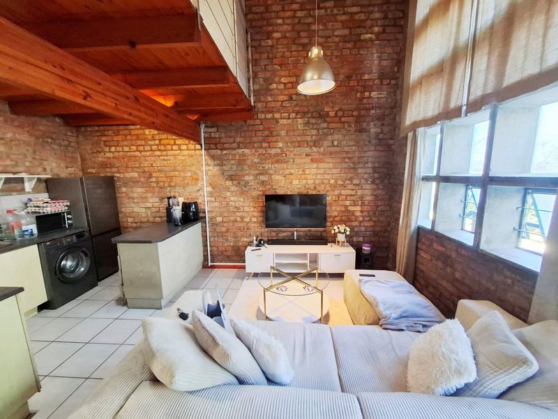 Open-plan living area featuring industrial-style exposed brick walls, timber ceiling beams, and a mezzanine level, creating a spacious and character-filled apartment interior.
