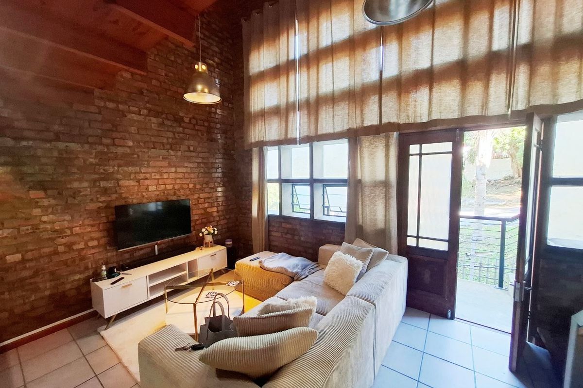 Spacious living room featuring exposed brick walls, high timber-beamed ceilings, tiled floors, and direct access to an outdoor balcony through a timber-framed glass door.