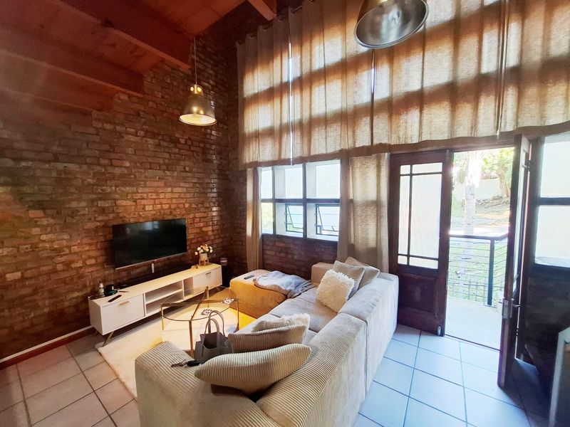 Spacious living room featuring exposed brick walls, high timber-beamed ceilings, tiled floors, and direct access to an outdoor balcony through a timber-framed glass door.