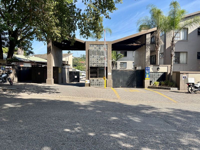 The entrance to the apartment complex features a secure gated access point with a boom gate and paved driveway.