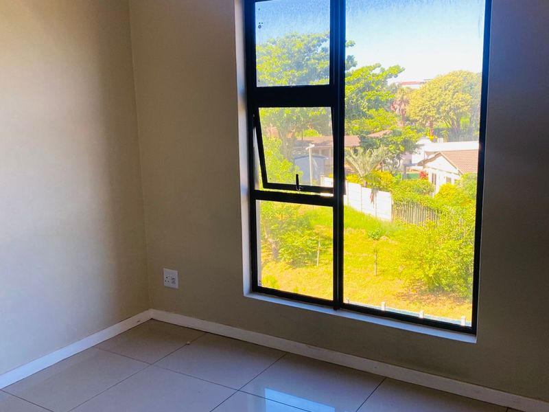 A bright, empty bedroom featuring neutral tiled flooring, a large aluminium-framed window, and a curtain rod, offering a pleasant view of the surrounding greenery.