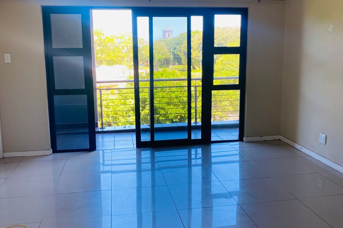 An empty, well-lit living area featuring polished tile flooring and large sliding glass doors that open onto a private balcony with green views.