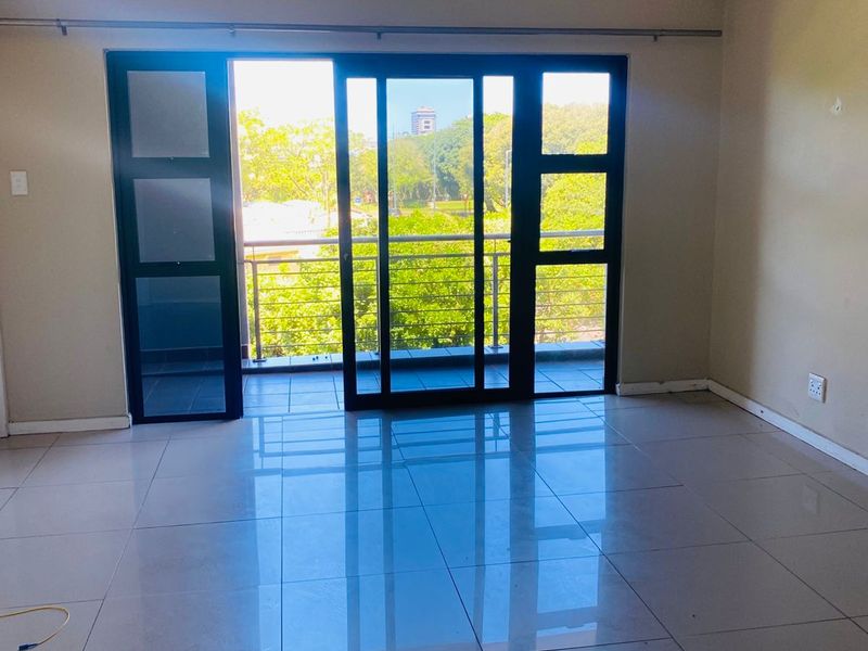An empty, well-lit living area featuring polished tile flooring and large sliding glass doors that open onto a private balcony with green views.