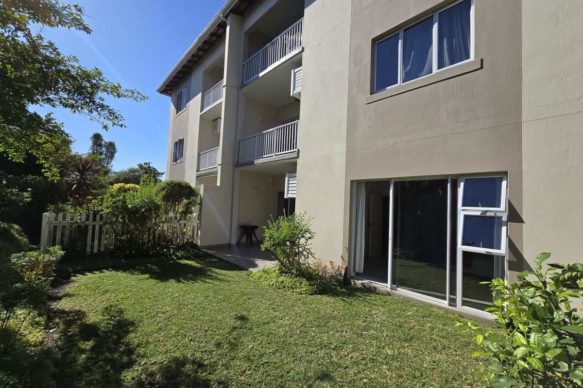 Rear view of the apartment building featuring private balconies, ground floor patio access with sliding doors, and a landscaped garden area.
