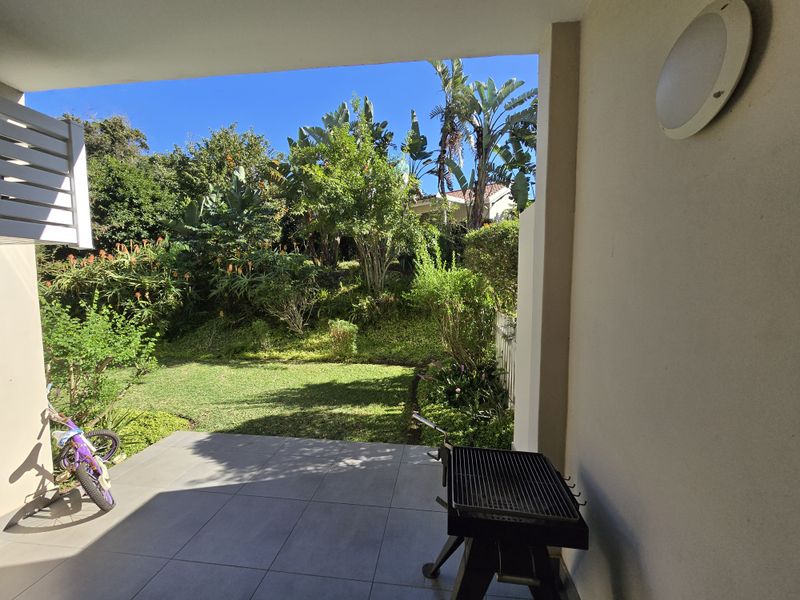 A covered patio area featuring tiled flooring and a wall-mounted light, overlooking a private garden space.