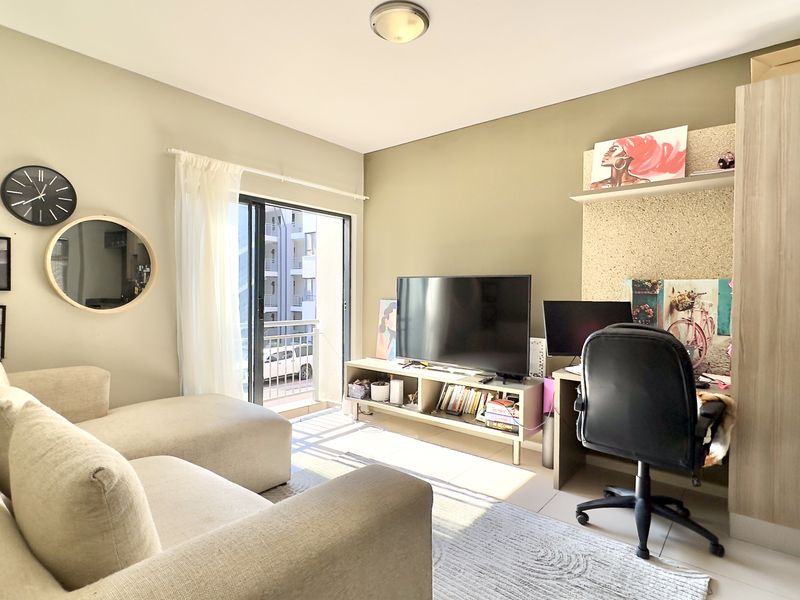 A well-lit living room featuring tiled flooring, a built-in desk area, and a sliding glass door leading to a balcony.