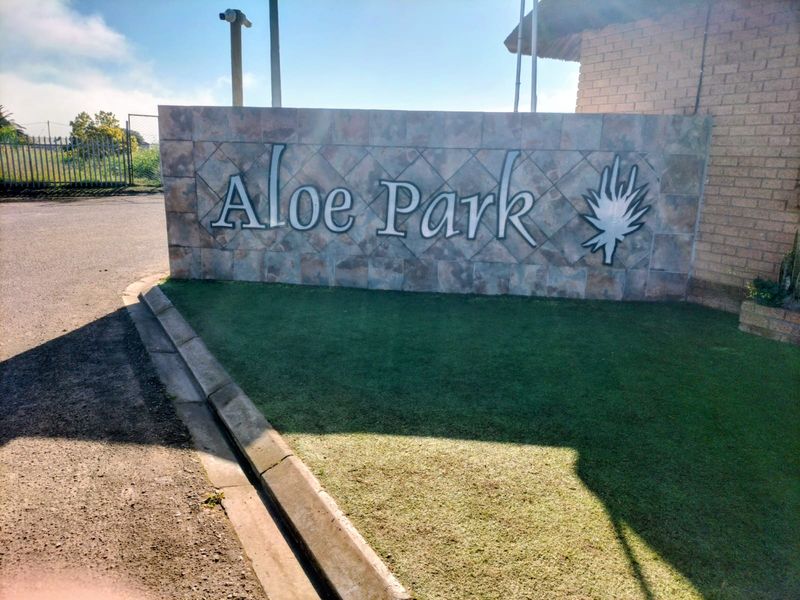 A clear view of the Aloe Park residential estate entrance signage wall, featuring stone cladding and adjacent paved driveway and lawn area.