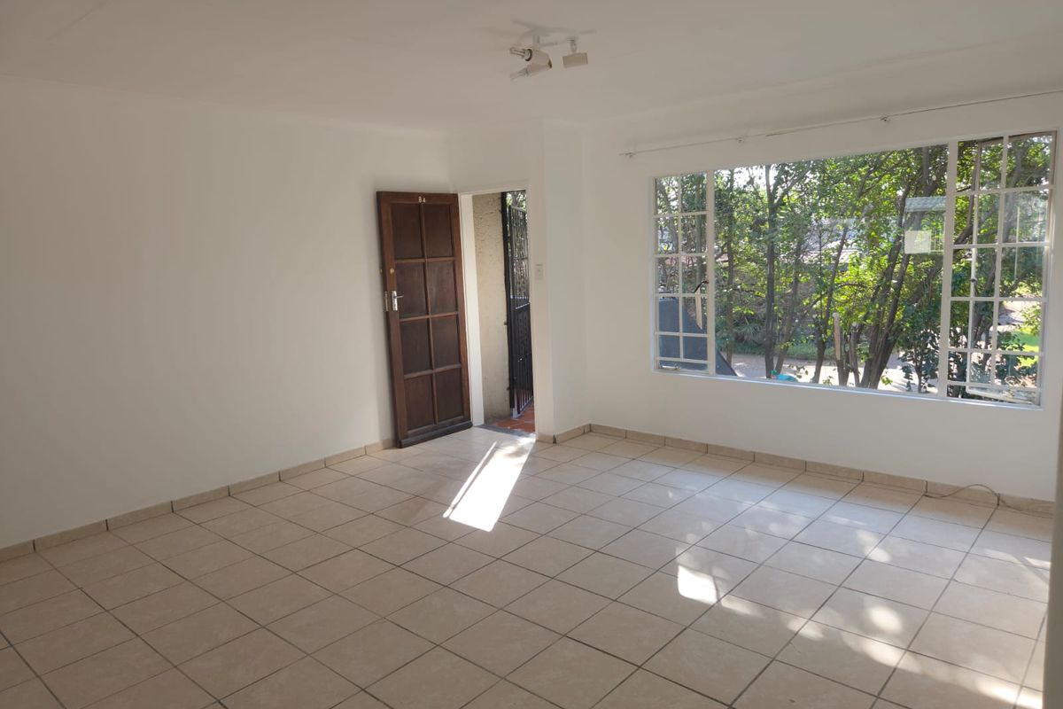 A bright, empty living room featuring neutral tiled flooring, a large window with garden views, and a timber entrance door.