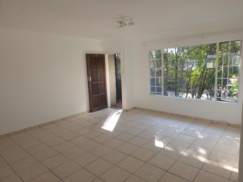 A bright, empty living room featuring neutral tiled flooring, a large window with garden views, and a timber entrance door.