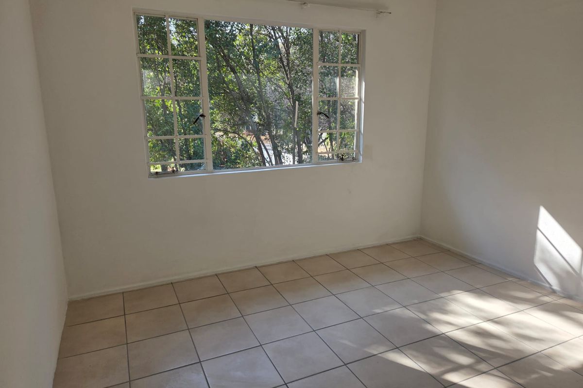 A bright, empty bedroom featuring neutral tiled flooring and a large multi-pane window overlooking mature trees.