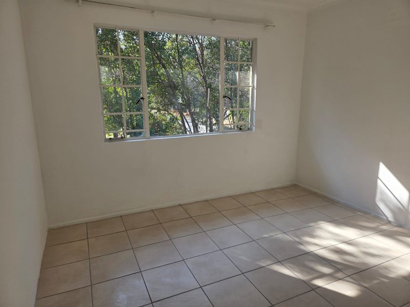 A bright, empty bedroom featuring neutral tiled flooring and a large multi-pane window overlooking mature trees.