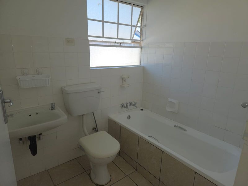 A functional bathroom featuring a built-in bathtub, wall-mounted basin, and toilet, finished with white wall tiles and neutral floor tiling.