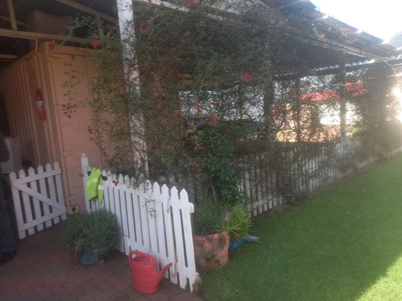A covered outdoor patio area featuring brick paving and a white picket fence, currently obscured by overgrown climbing plants.