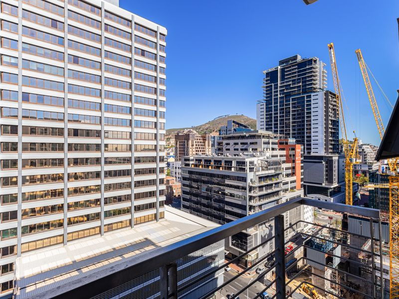 An elevated urban view from a balcony overlooking surrounding high-rise buildings and city infrastructure under a clear blue sky.
