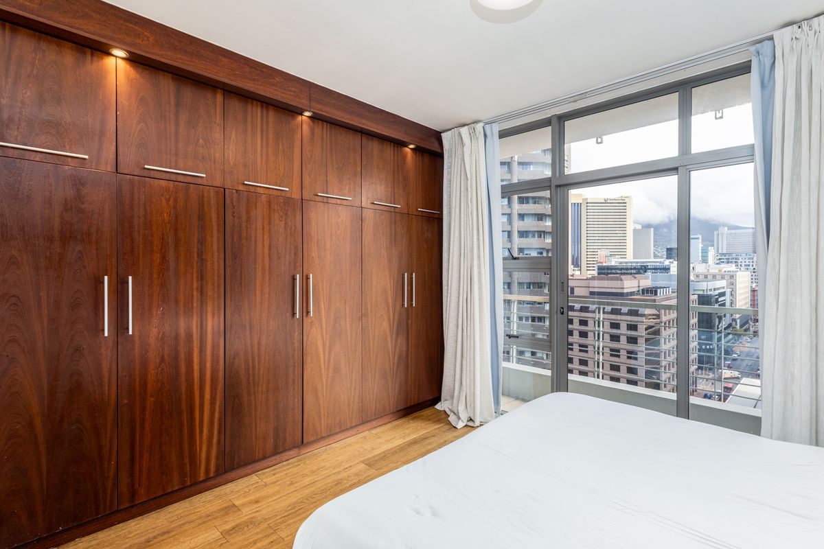 A well-maintained bedroom featuring extensive built-in wooden cabinetry, laminate flooring, and large floor-to-ceiling windows offering an urban city view.