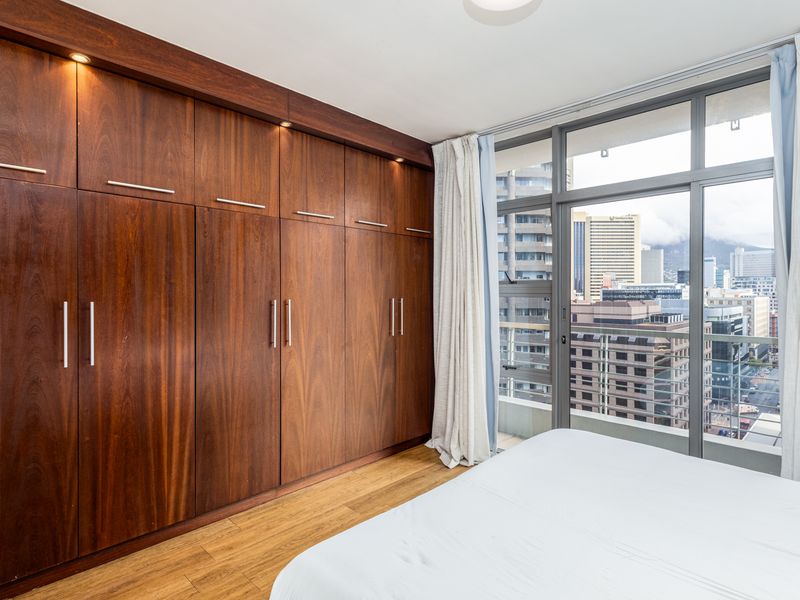 A well-maintained bedroom featuring extensive built-in wooden cabinetry, laminate flooring, and large floor-to-ceiling windows offering an urban city view.