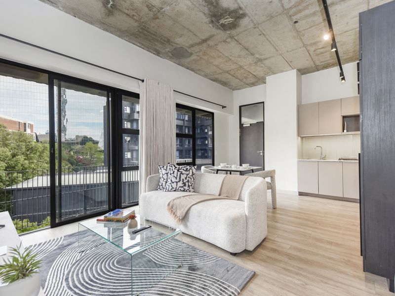 Modern open-plan living area featuring an exposed concrete ceiling, sleek kitchen cabinetry, and large sliding glass doors leading to a balcony.
