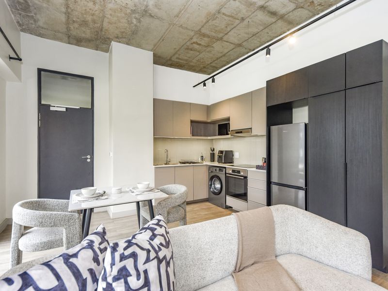 Modern open-plan apartment featuring an exposed concrete ceiling, sleek built-in kitchen cabinetry, integrated appliances, and contemporary track lighting.