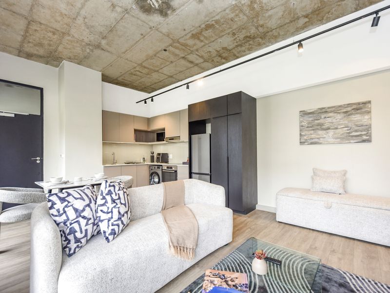 Modern open-plan living area featuring an exposed concrete ceiling, track lighting, and a sleek, compact kitchen with integrated appliances and laminate flooring.
