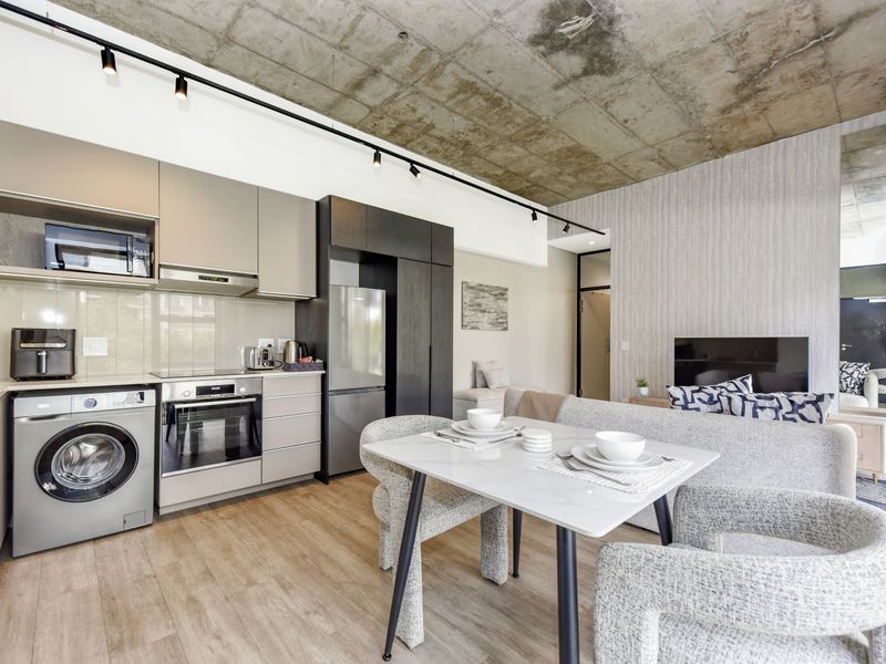 Modern open-plan living area featuring an exposed concrete ceiling, track lighting, and a sleek, well-equipped kitchen with integrated appliances and laminate flooring.