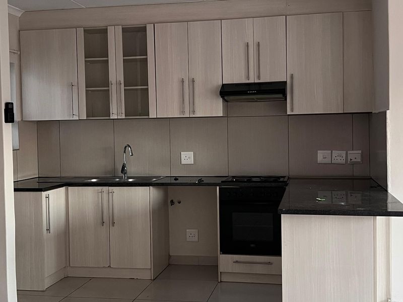 A clean, modern kitchen featuring light wood-finish cabinetry, dark granite countertops, built-in oven, extractor fan, and neutral floor tiles.
