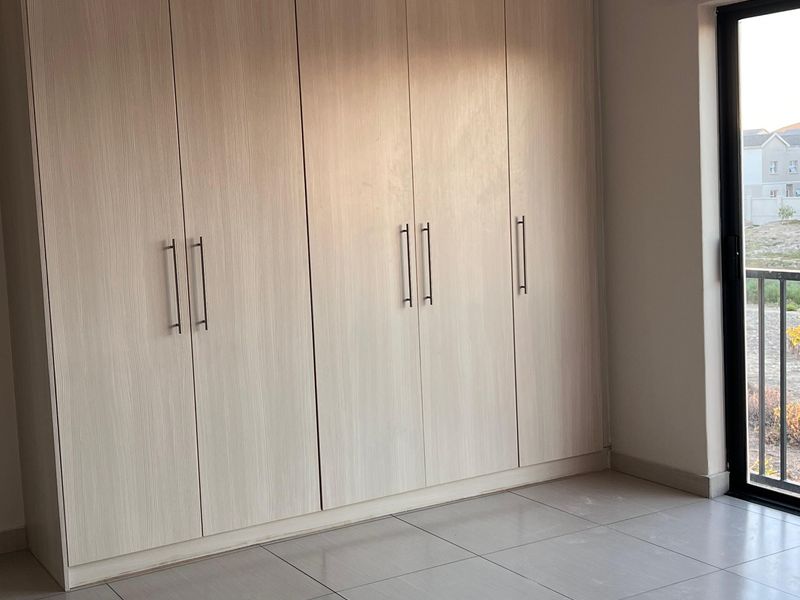A bright, empty bedroom featuring modern light wood built-in cupboards, neutral tiled flooring, and a sliding door leading to an outdoor area.