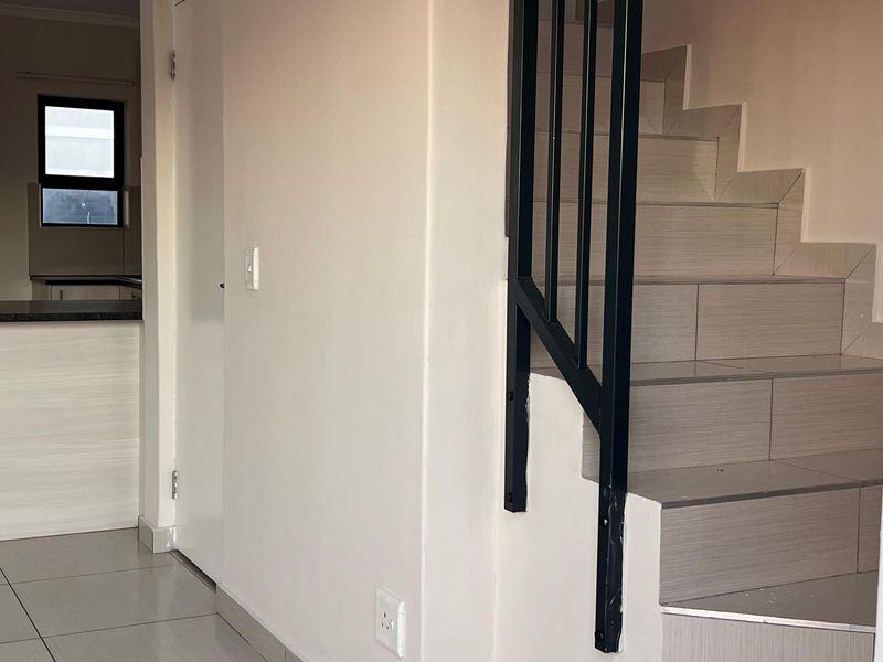 A bright, modern hallway featuring a tiled staircase with a sleek black metal balustrade and neutral ceramic floor tiles.