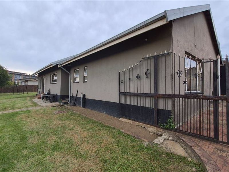 Rear view of the property featuring a paved driveway, wrought iron gate, and a spacious lawn area with plastered exterior walls.