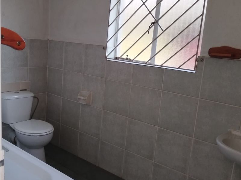 A functional bathroom featuring a bathtub, pedestal basin, and toilet with neutral wall and floor tiling, secured by window bars.