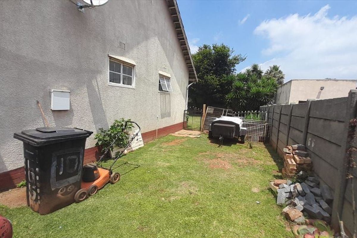 A side yard area featuring a lawn, precast concrete boundary wall, and metal gate access, showing signs of wear on the exterior wall base.