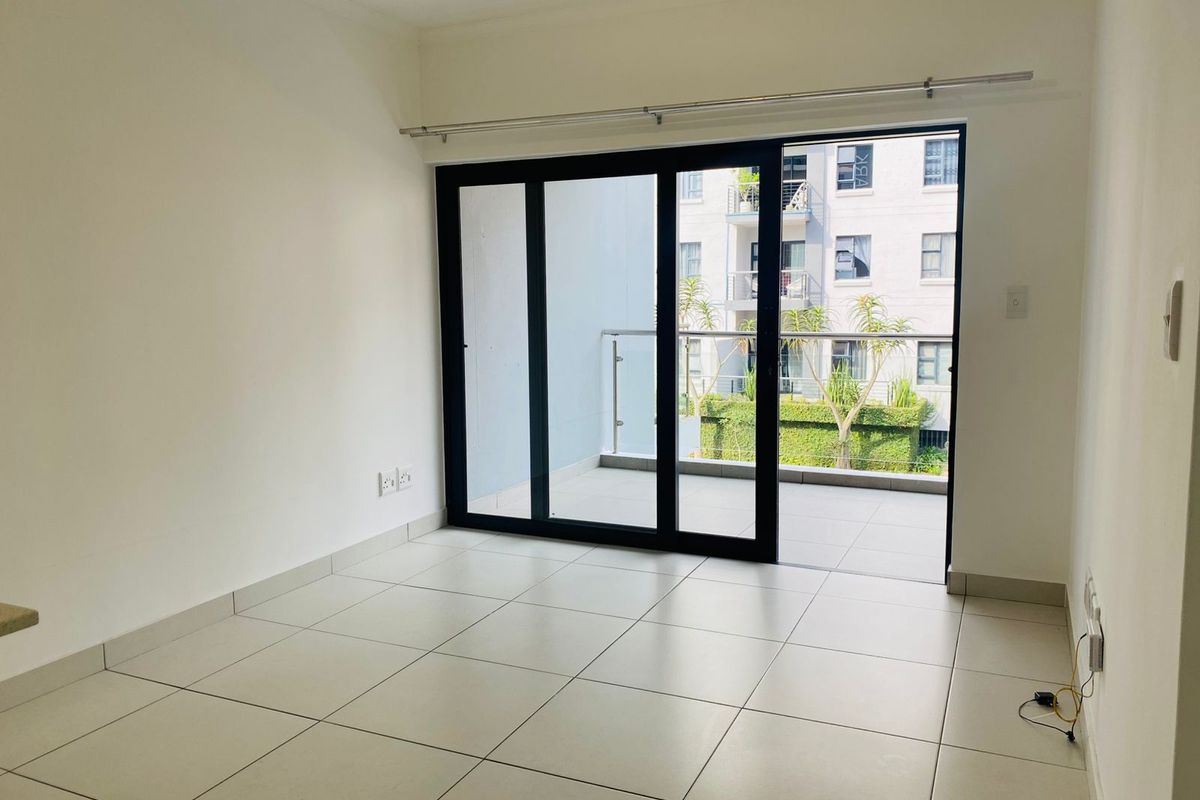 An empty, well-lit living area featuring neutral tiled flooring and large sliding glass doors that open onto a private balcony.