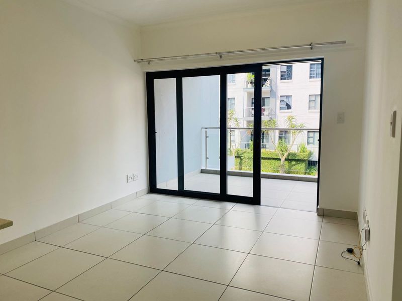 An empty, well-lit living area featuring neutral tiled flooring and large sliding glass doors that open onto a private balcony.