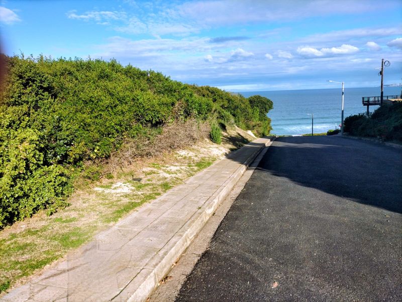 A street view showing the asphalt road and concrete sidewalk leading towards the coastline with ocean views.