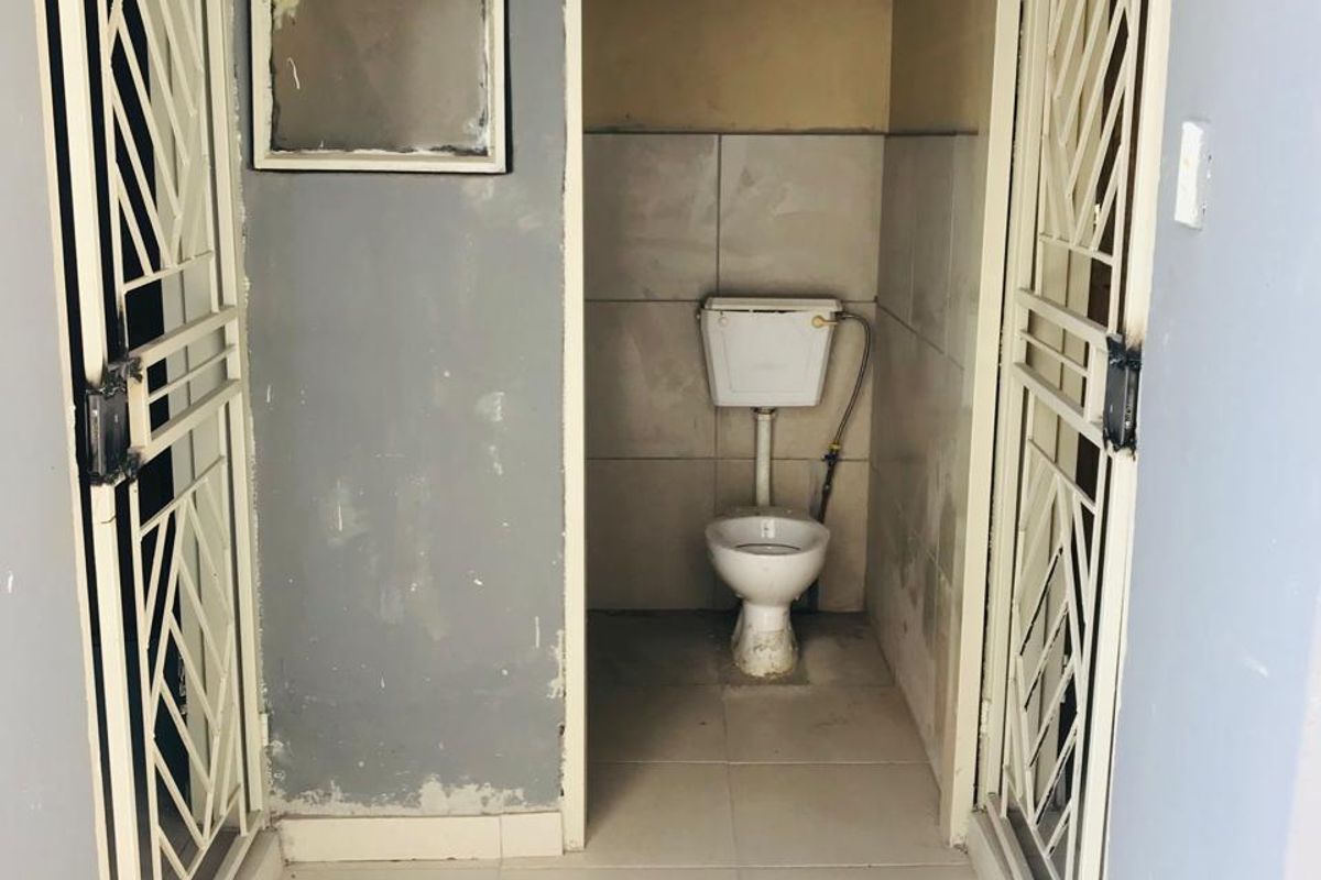 A small, basic bathroom featuring a toilet, tiled flooring, and security gates on the entryways. The space requires cosmetic updates and wall repairs.