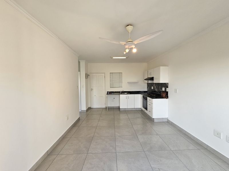 An open-plan kitchen and living area featuring tiled floors, white cabinetry, black granite countertops, and a ceiling fan in a clean, neutral space.