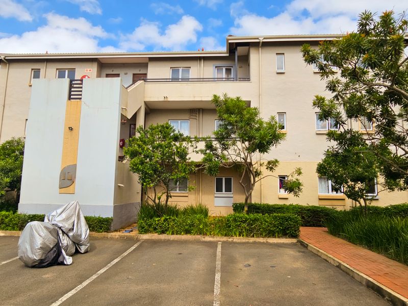 Front elevation of a multi-story apartment complex featuring paved parking, landscaped garden beds, and accessible stairwell and balcony areas.
