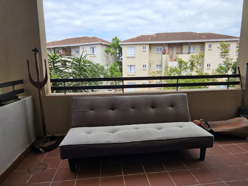 A covered balcony featuring tiled flooring and a metal balustrade, offering an outdoor space with views of the surrounding apartment complex.