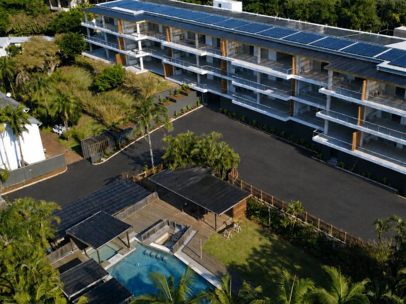 Aerial view of a modern apartment complex featuring solar panels, a swimming pool with timber decking, and landscaped grounds.