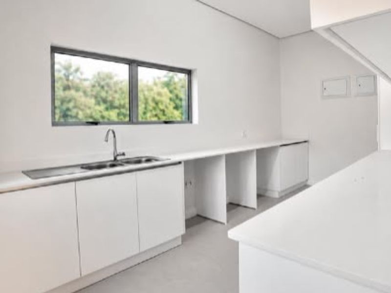 A clean, modern scullery featuring white built-in cabinetry, stone countertops, a stainless steel sink, and a large window providing natural light.