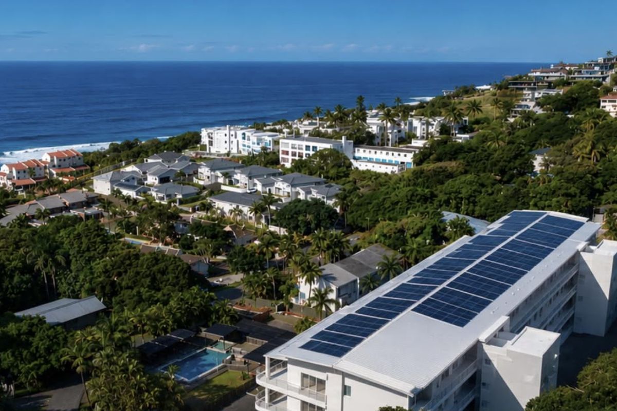 Aerial view of a coastal apartment complex featuring rooftop solar panels, a swimming pool, and surrounding lush greenery with ocean views.