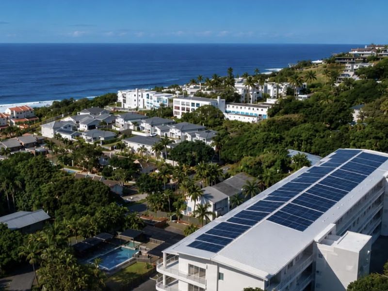 Aerial view of a coastal apartment complex featuring rooftop solar panels, a swimming pool, and surrounding lush greenery with ocean views.