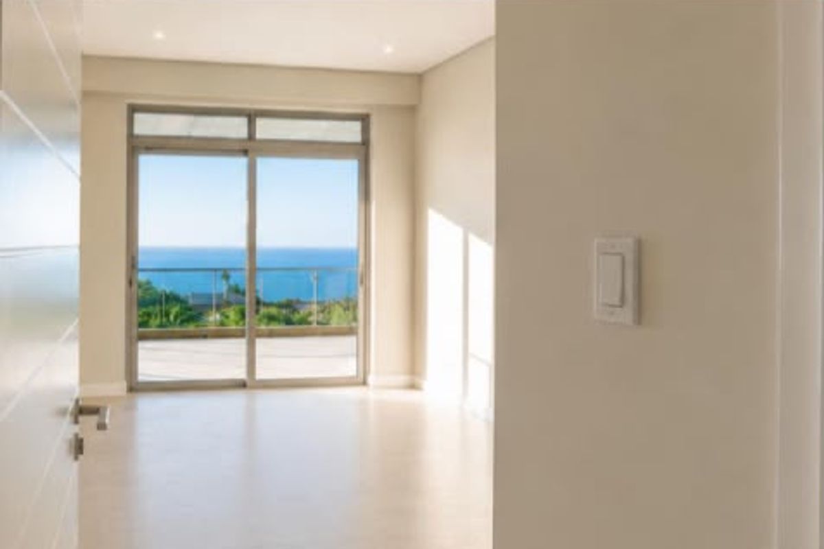 A bright, empty bedroom featuring neutral finishes, recessed lighting, and a sliding glass door opening onto a balcony with scenic ocean views.