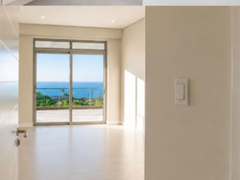 A bright, empty bedroom featuring neutral finishes, recessed lighting, and a sliding glass door opening onto a balcony with scenic ocean views.