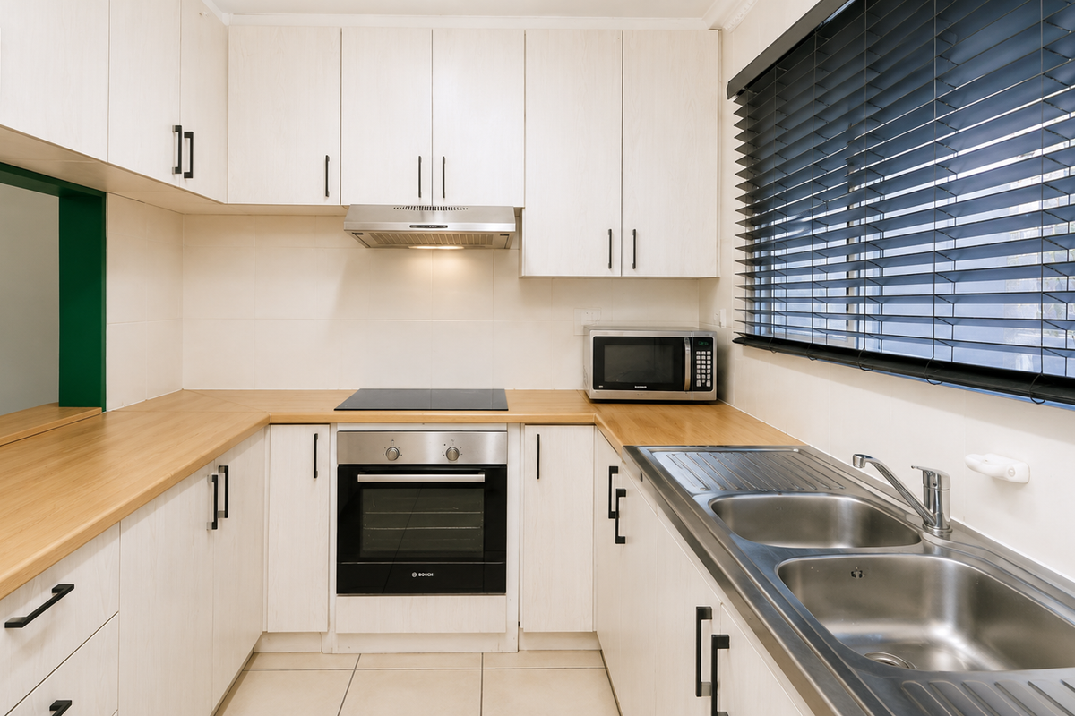 A well-maintained kitchen featuring light wood-grain cabinetry, laminate countertops, integrated oven and hob, and a double stainless steel sink with venetian blinds.